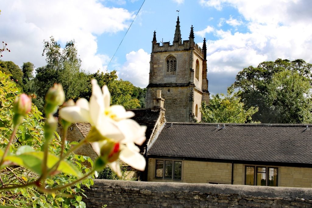 Church tower rising above the stone rooftops of Castle Combe, framed by flowers and green trees