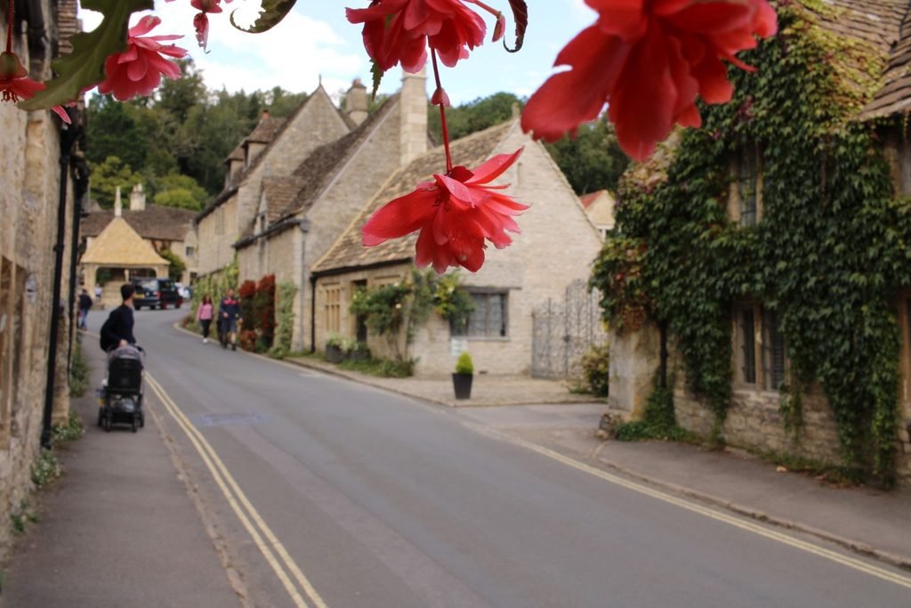 Quiet Castle Combe street lined with historic stone houses and red flowers hanging in the foreground