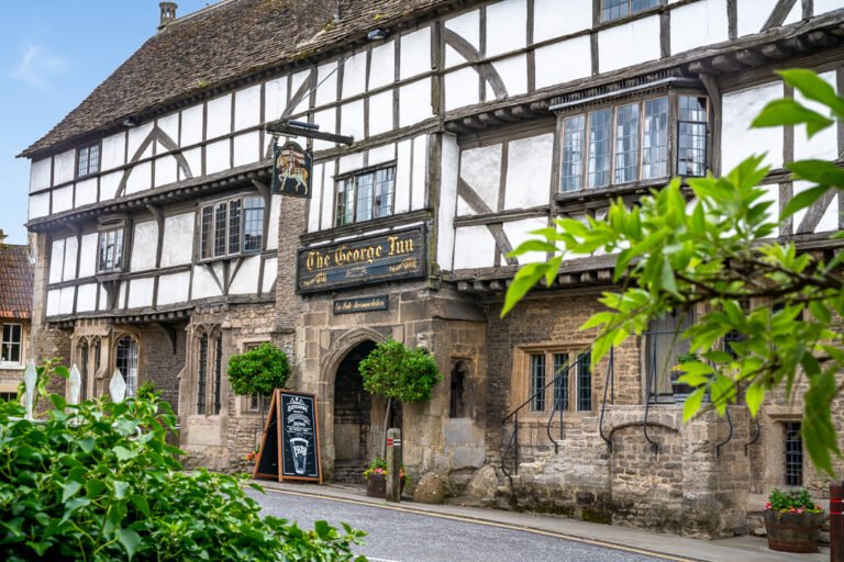 The historic George Inn in Norton St Philip, Somerset, with its long timber-framed upper floor, stone lower walls, old pub sign, and warmly lit windows on a damp English afternoon
