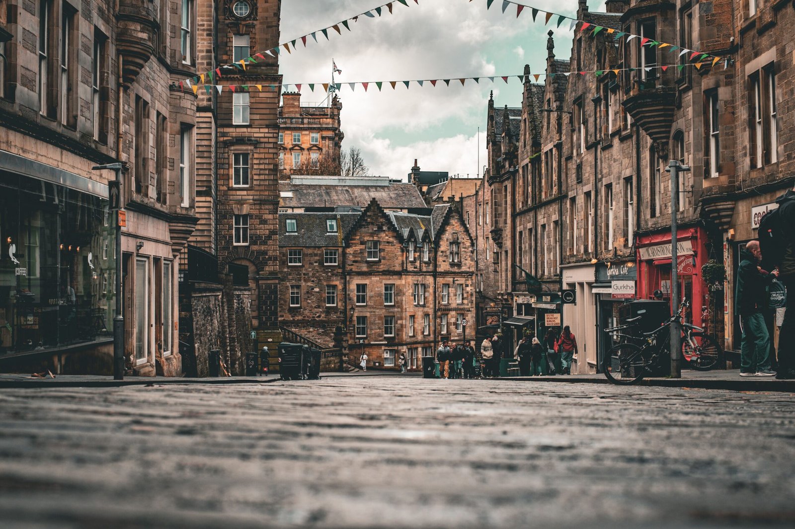 Stone buildings, colorful bunting, and pedestrians along a cobbled Edinburgh street beneath a cloudy sky